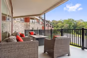 Covered outdoor patio area with wicker furniture including a sofa and chairs with red cushions, a fire pit table in the center, and a black railing overlooking greenery and a stone building wall.