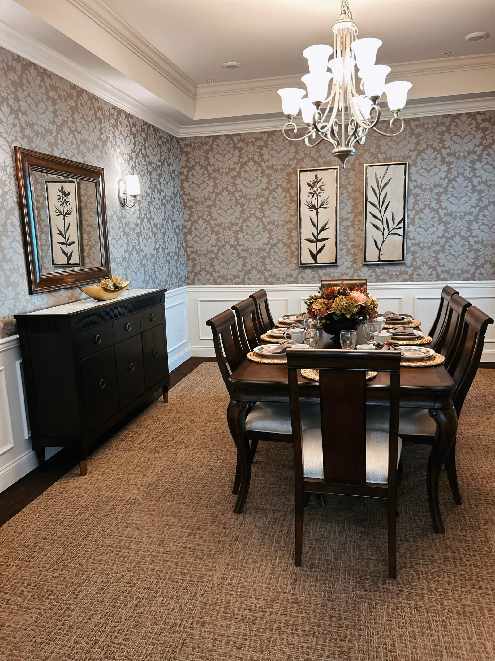 Formal dining room with a wooden table set for eight, a floral centerpiece, chandelier, sideboard, and patterned wallpaper.