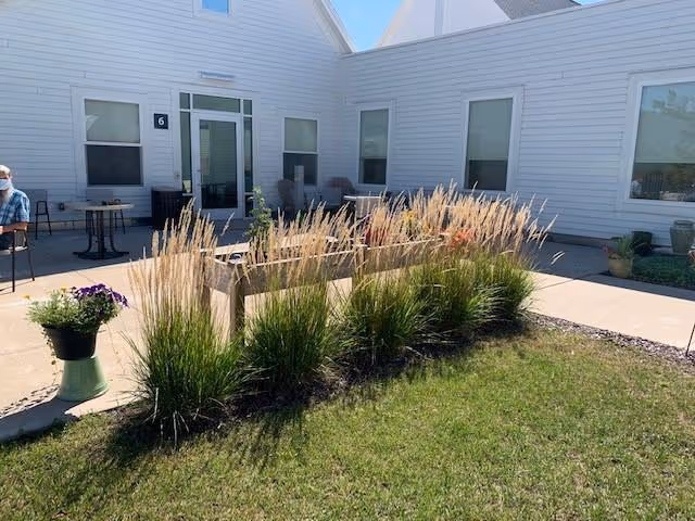 Outdoor courtyard area at Juliette Manor featuring a grassy lawn, ornamental grasses planted in a row, potted plants, and a paved patio with tables and chairs. The building exterior is white with several windows and a door marked with the number 6. A person wearing a mask is seated at one of the tables.