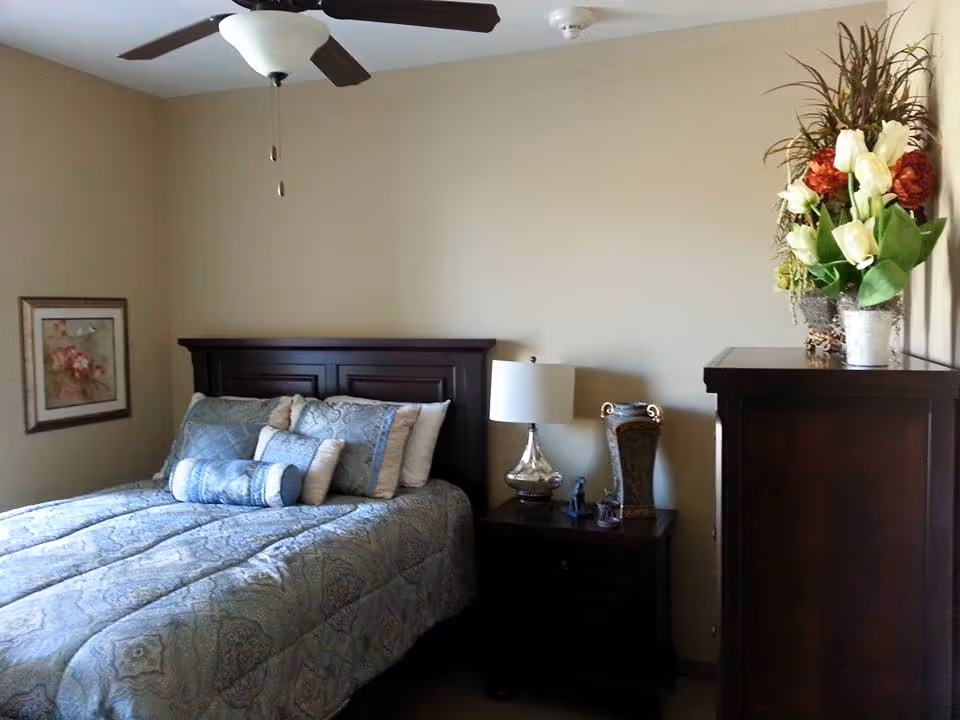 Bedroom with a patterned bed, wooden headboard, nightstand with lamp, tall dresser, ceiling fan, and a vase of flowers.