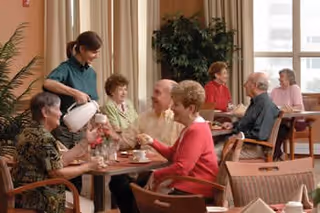 A group of elderly people sitting around tables in a dining area, with a caregiver pouring a beverage for one of the residents. The room has large windows, plants, and light-colored curtains, creating a warm and welcoming atmosphere.