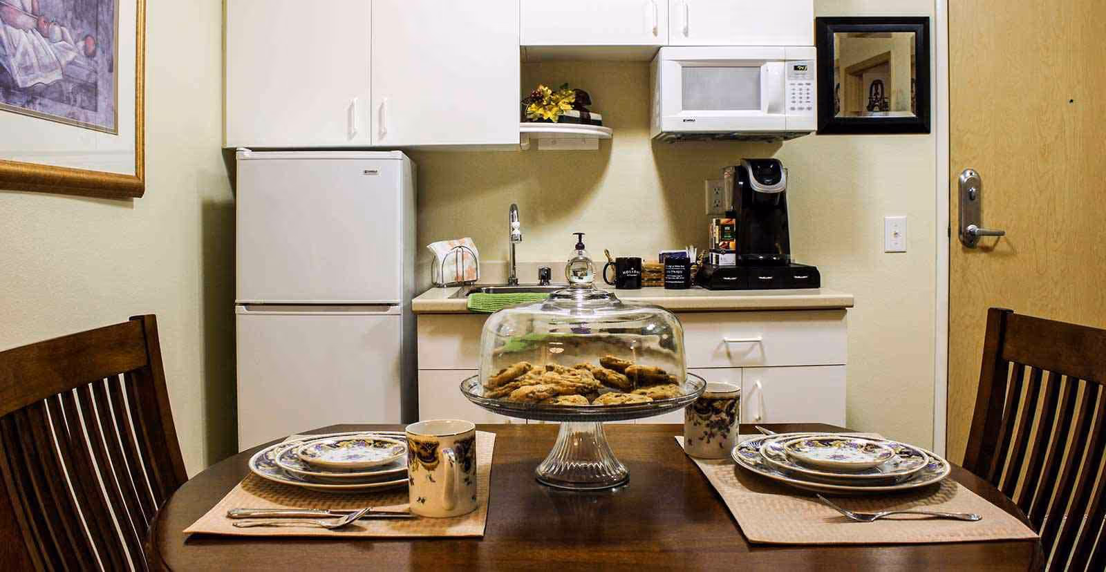 A small kitchen area with white cabinets, a refrigerator, microwave, and coffee maker. In the foreground, a wooden dining table is set for two with plates, mugs, and utensils. A glass cake stand with cookies is centered on the table.