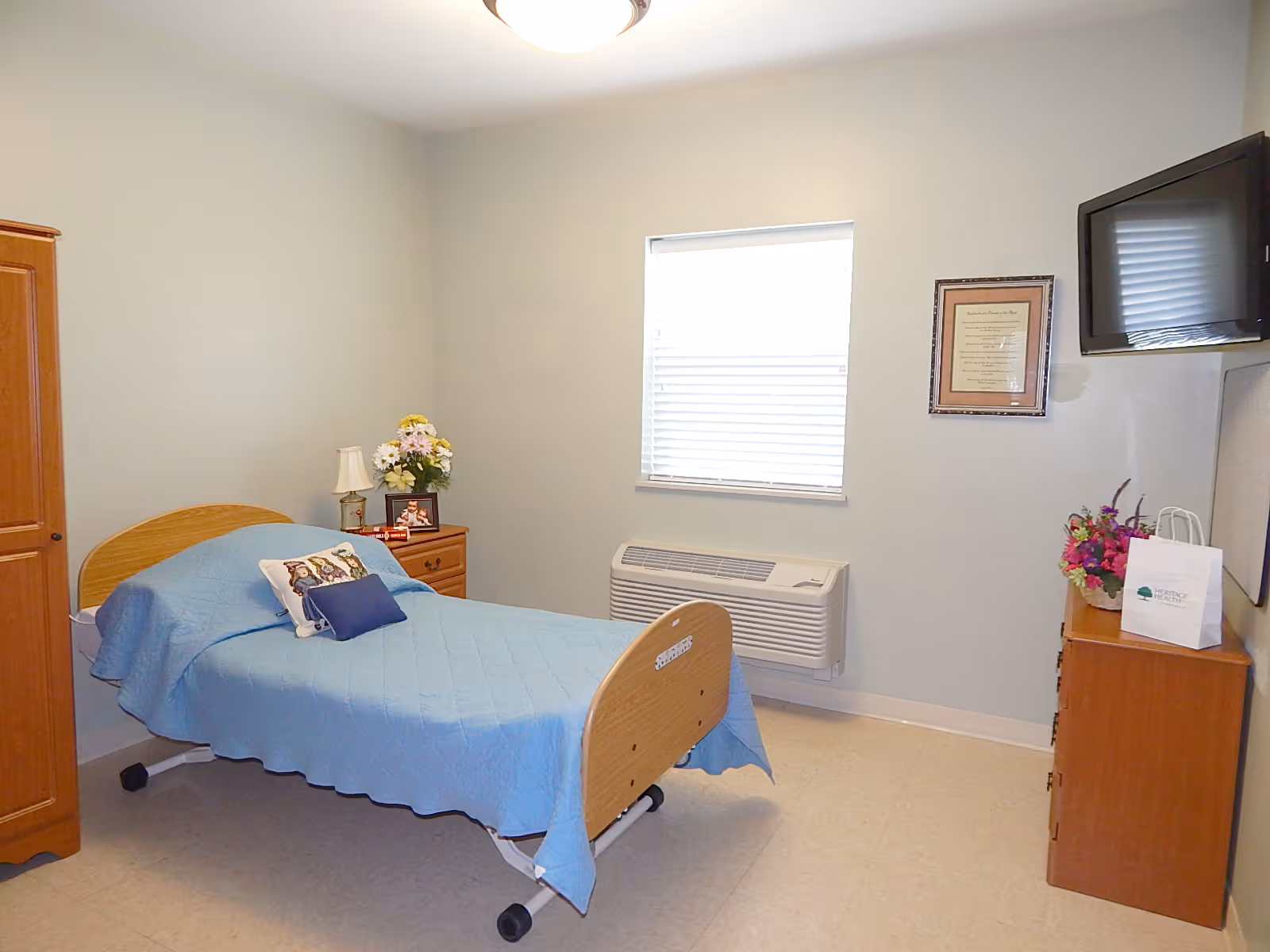 A simple, clean bedroom with a single bed covered in a blue quilt. The bed has a pillow with a dog design and a smaller blue pillow. There is a wooden nightstand with a lamp, flowers, and a framed photo. A wooden dresser with a bouquet of flowers and a white paper bag is on the right side. A flat-screen TV is mounted on the wall above the dresser. A window with blinds and an air conditioning unit below it are centered on the back wall. The walls are painted light blue, and the floor is light-colored linoleum.