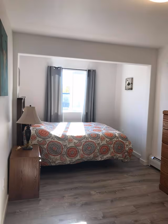 A bedroom with a bed covered in a patterned bedspread featuring circular designs in red, gray, and beige. There is a wooden nightstand with a lamp on the left side of the bed and a wooden dresser on the right side of the room. A window with gray curtains is centered on the back wall, allowing natural light to enter the room. The floor is a light wood laminate, and the walls are painted white with minimal decorations.