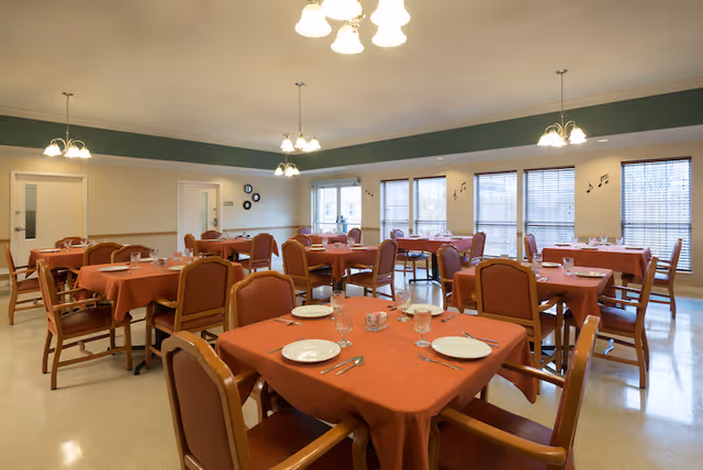 A dining room with multiple tables covered in orange tablecloths, each set with plates, glasses, and silverware. The room has beige walls, large windows with blinds, and ceiling lights. Chairs with wooden frames and orange cushions surround the tables.
