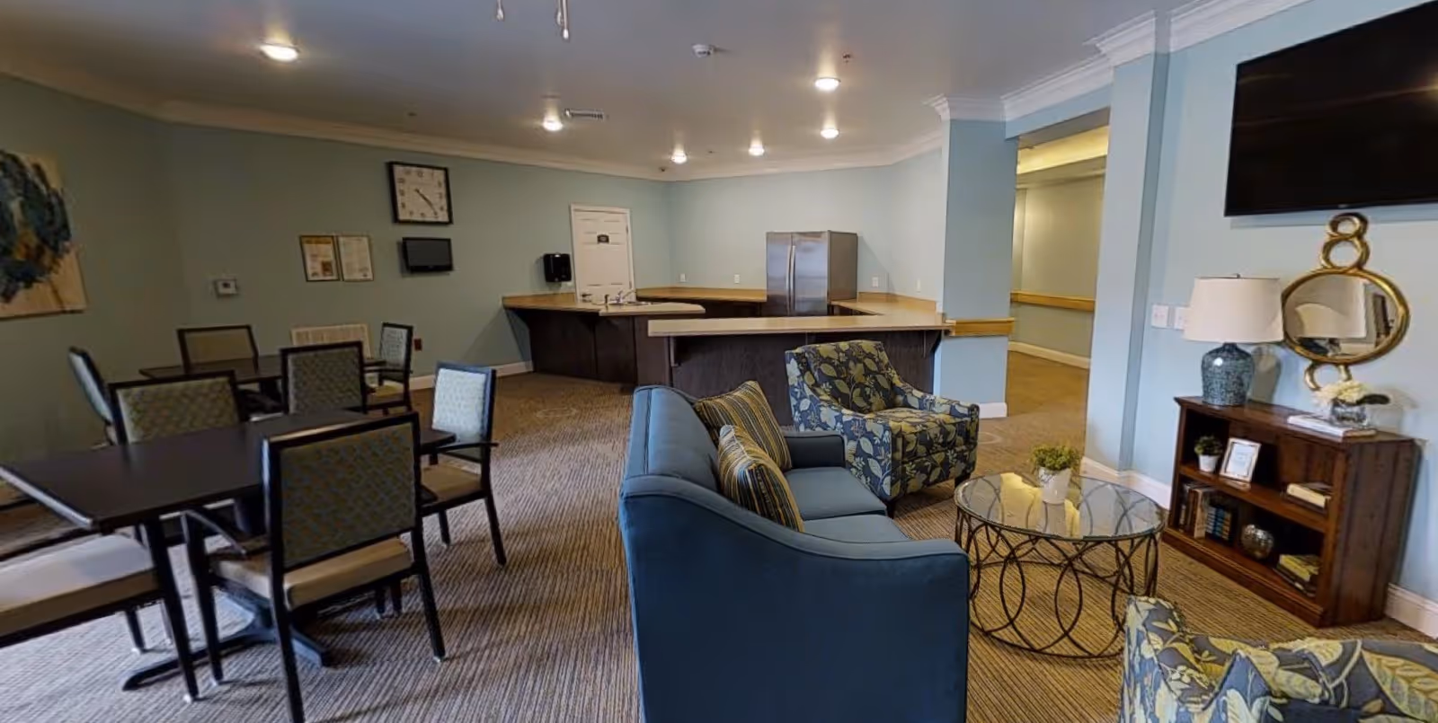 A communal living area with blue walls featuring a seating area with a blue sofa and two patterned armchairs around a glass coffee table. There is a wooden cabinet with a lamp, mirror, and decorative items beneath a wall-mounted TV. In the background, there is a kitchenette with a refrigerator, sink, and counter space, and a dining area with tables and chairs.