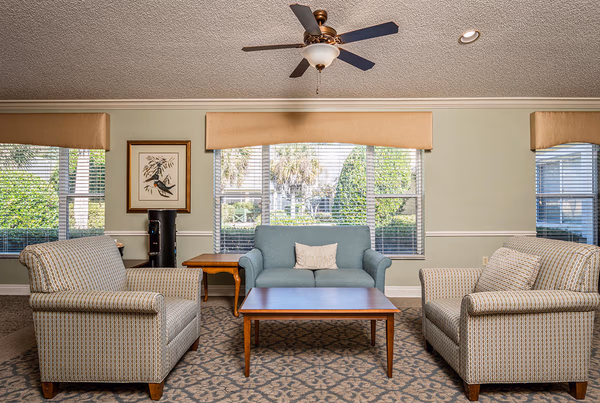 A cozy living room area with two patterned armchairs facing a blue loveseat with a decorative pillow. A wooden coffee table is centered between the seating. Large windows with beige valances let in natural light and show greenery outside. A ceiling fan with a light fixture hangs from the textured ceiling. A framed bird artwork is on the wall next to a black water cooler.