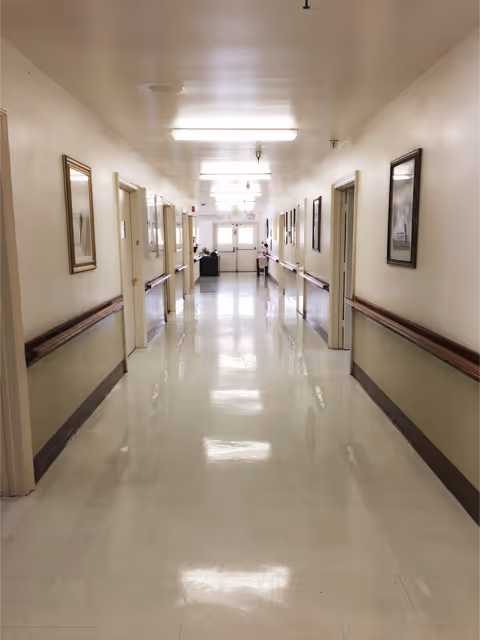 Long, brightly lit interior hallway in a care facility with handrails, doors, and framed pictures along the walls.