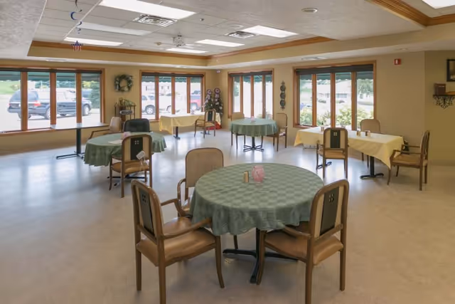 A spacious dining room with several round and rectangular tables covered with green and yellow tablecloths. Each table is surrounded by wooden chairs with cushioned seats. Large windows line the walls, allowing natural light to fill the room and providing a view of parked cars outside. The ceiling has recessed lighting and wooden trim accents.