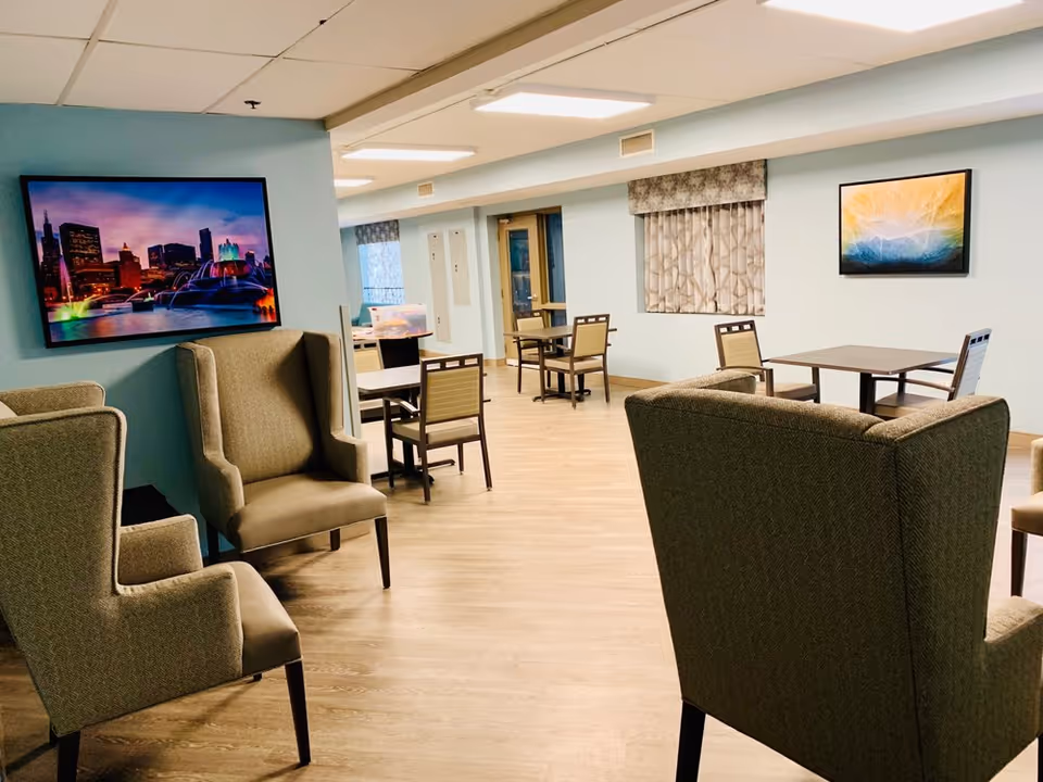 Bright common lounge and dining area with armchairs, tables, wall art, and a television.