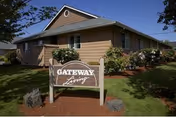Exterior view of a single-story building with beige siding and a gabled roof, surrounded by green grass, bushes, and trees. In front of the building is a wooden sign that reads 'Gateway Living'.