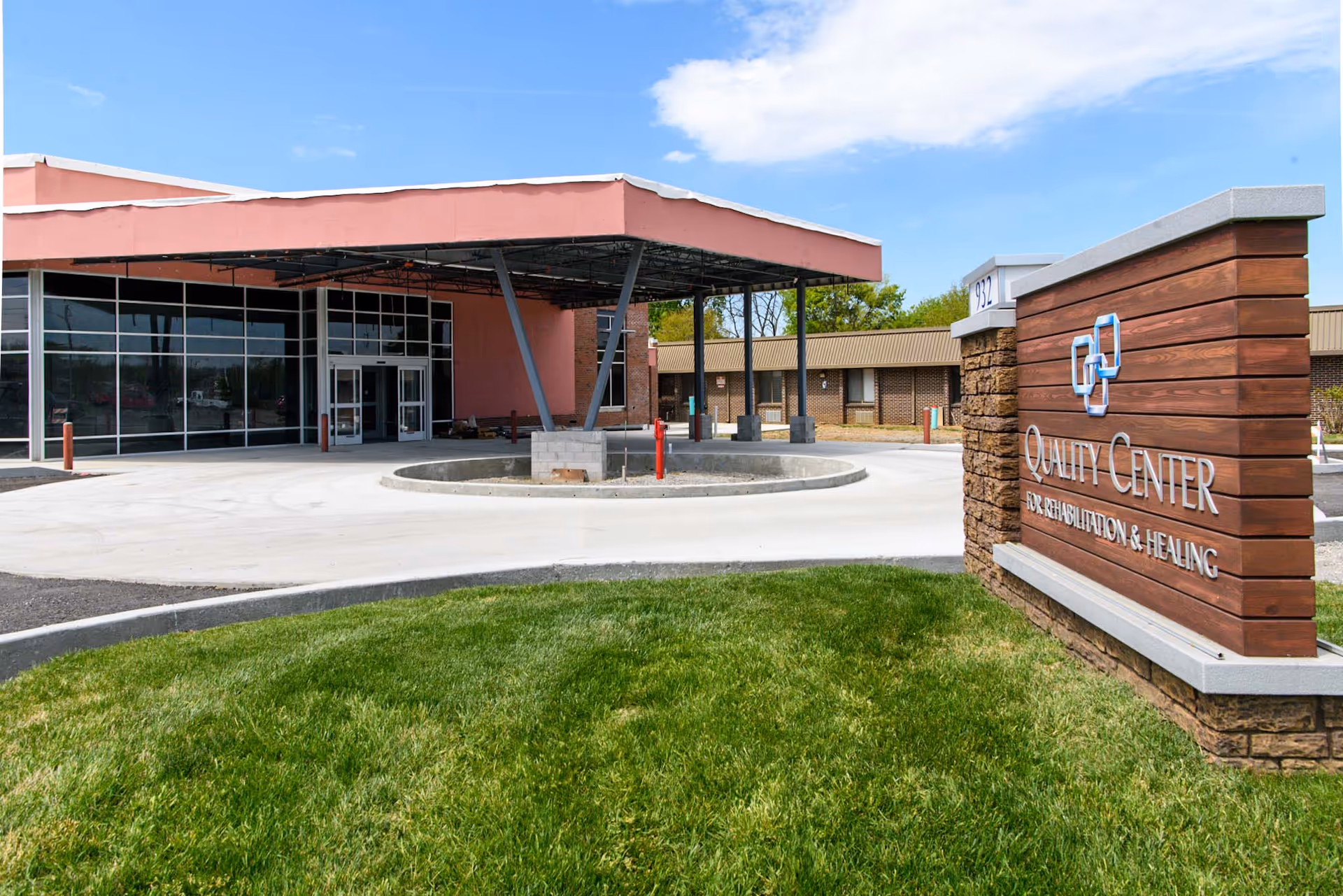 Exterior view of the Quality Center for Rehabilitation and Healing building with a covered entrance, large glass windows, and a sign displaying the facility's name on a wooden and stone base, surrounded by green grass under a blue sky.