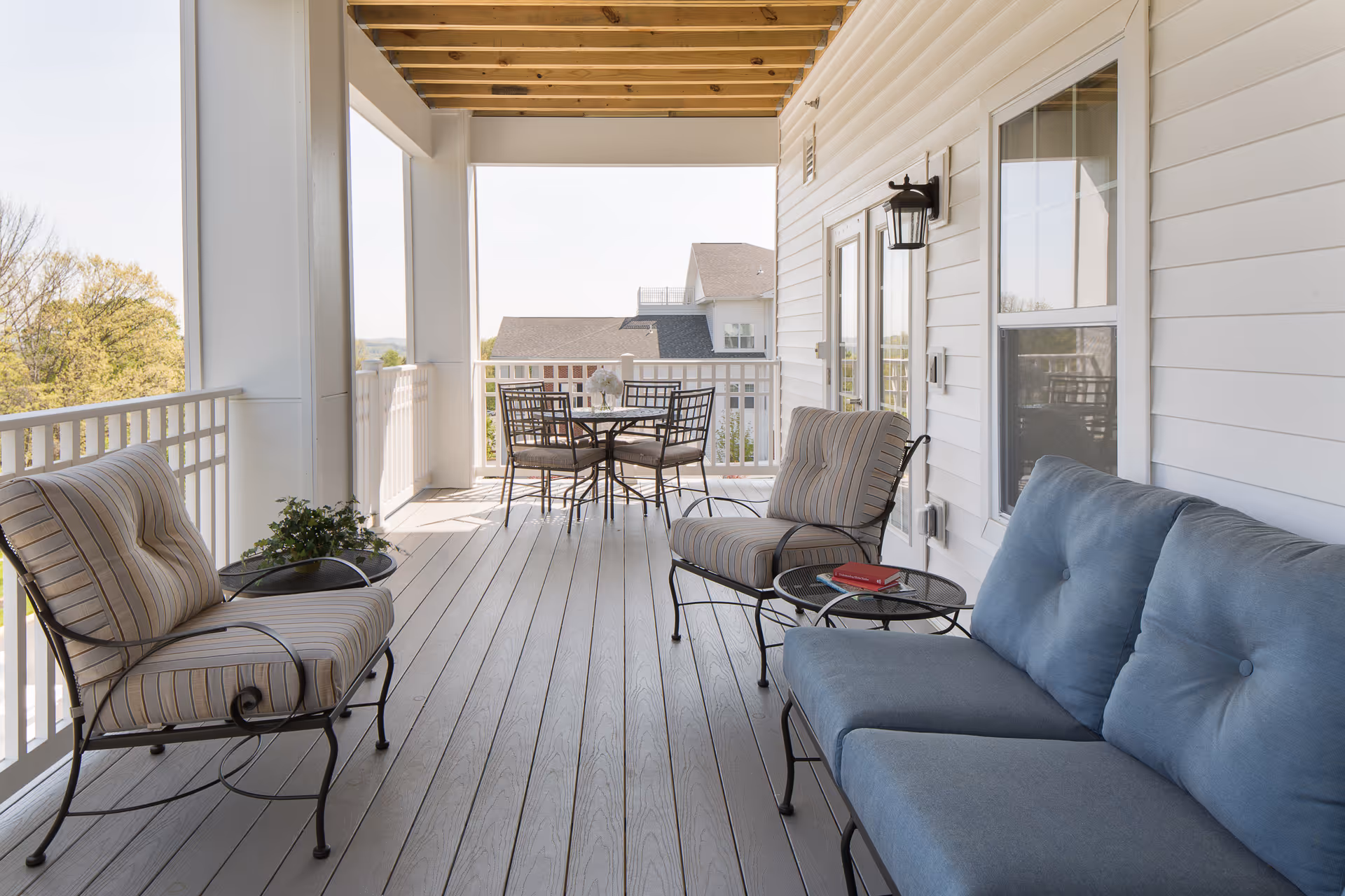 Covered outdoor porch with cushioned seating including a blue sofa and two striped armchairs, a small round table with a red book, and a dining table with four chairs in the background. The porch overlooks trees and neighboring houses.