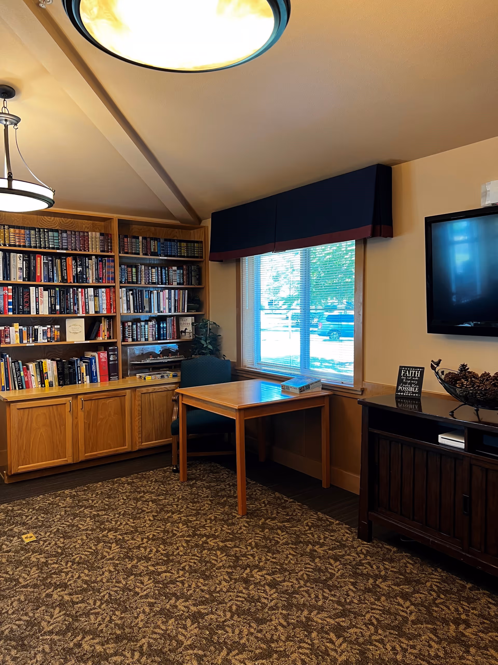 A cozy room with a wooden bookshelf filled with books, a wooden table with a chair, a window with blinds and a dark valance, a TV mounted on the wall, and a cabinet with decorative items including a bowl of pinecones and a small sign.