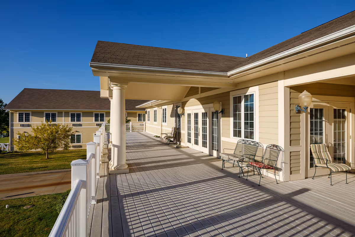 A spacious outdoor balcony area of a senior living facility with beige siding, white columns, and several metal chairs with cushions. The balcony overlooks a grassy area with a tree and another building in the background under a clear blue sky.