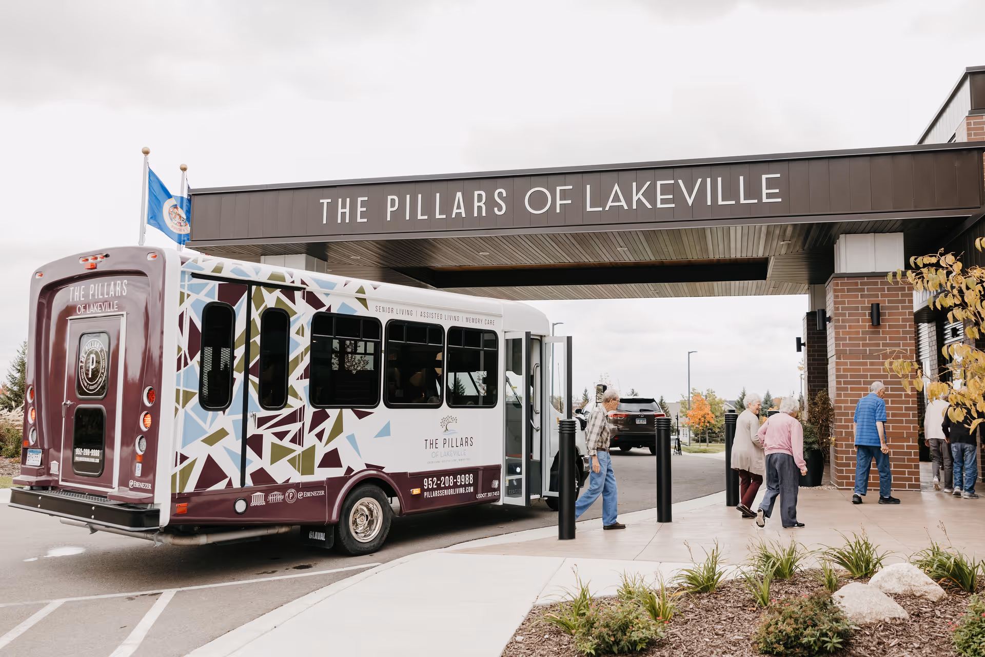 A shuttle bus with The Pillars of Lakeville branding is parked under a covered entrance of the facility. Several elderly people are walking towards the building entrance. The area is landscaped with plants and small rocks, and the sky is overcast.