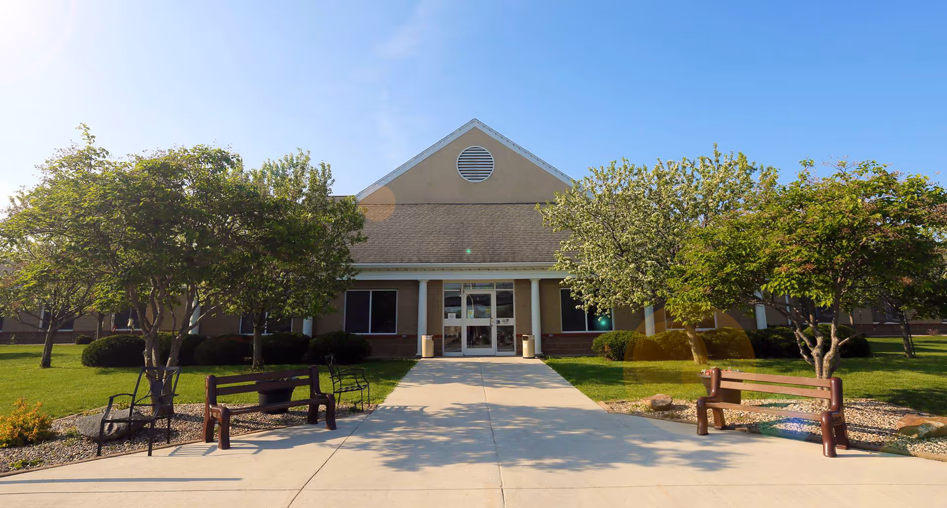 Front exterior view of a single-story building with a peaked roof, flanked by green trees and bushes. There is a concrete walkway leading to the entrance with glass double doors. Benches and chairs are placed on either side of the walkway.