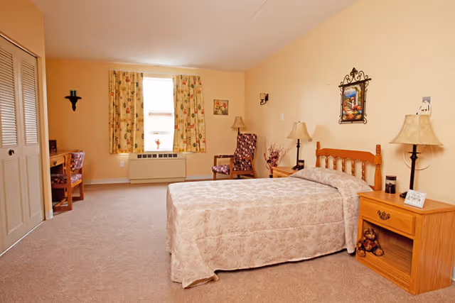 A neatly furnished senior living bedroom with a single bed, wooden nightstand and chair, a desk by the window, and floral curtains.