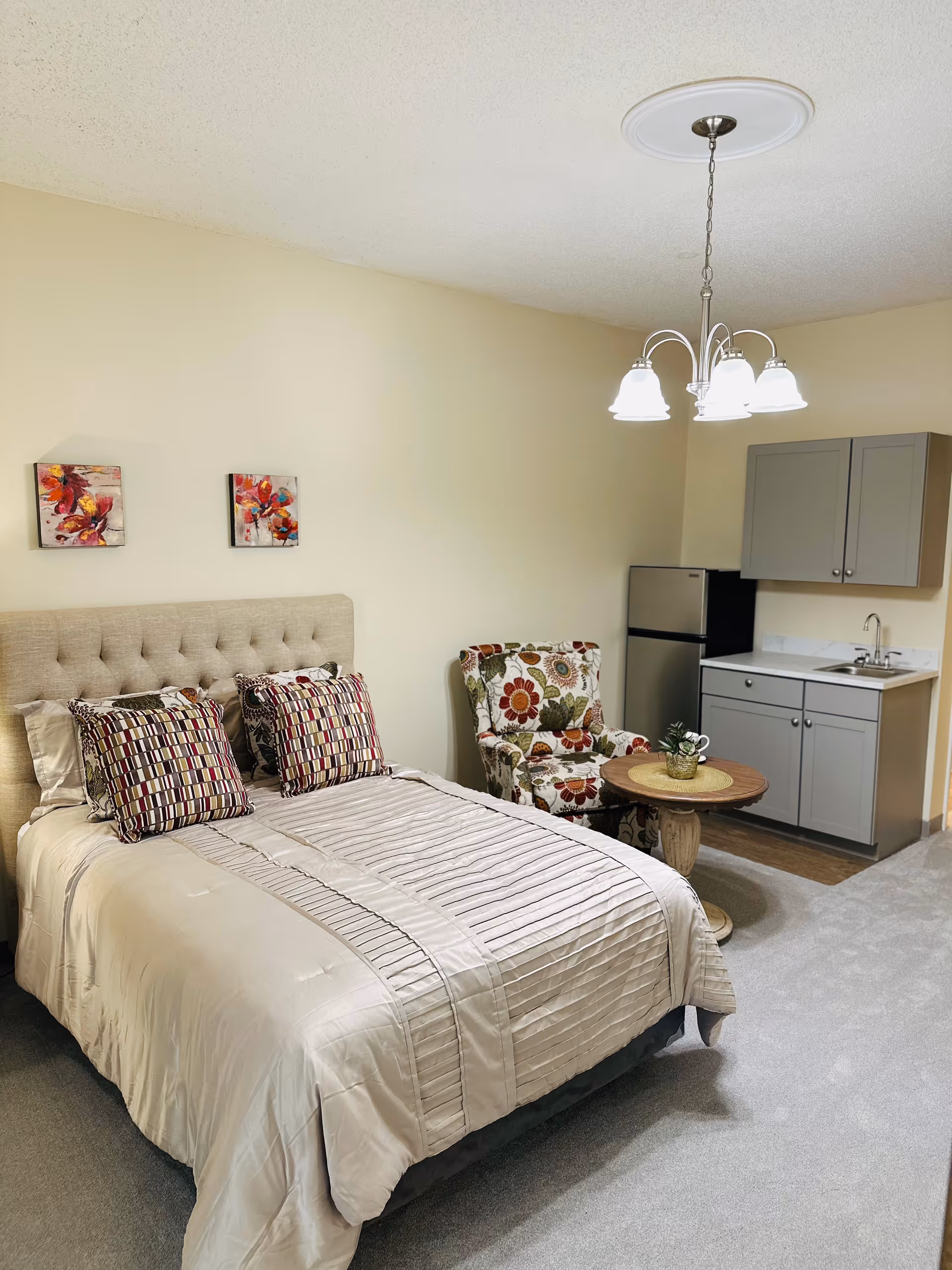 A senior living facility room featuring a neatly made bed with a beige tufted headboard and patterned pillows, a floral upholstered armchair next to a small round wooden table with a plant on it, and a kitchenette area with gray cabinets, a small refrigerator, and a sink. The room has beige walls and carpeted flooring, with a ceiling light fixture above.