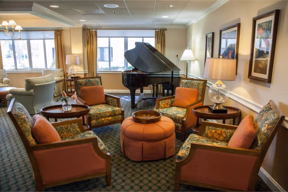 A cozy living room area with four floral-patterned armchairs with orange cushions arranged around a round orange ottoman. There are two round wooden side tables with lamps and a small vase with flowers. In the background, a black grand piano is positioned near windows with beige curtains. The room has carpeted flooring and framed artwork on the walls.