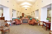 A spacious and well-lit common area in a nursing home featuring multiple armchairs arranged around a central fireplace. The room has high ceilings with recessed lighting, beige walls, and carpeted floors. There are side tables with lamps and plants, and a decorative wall clock is visible in the background.