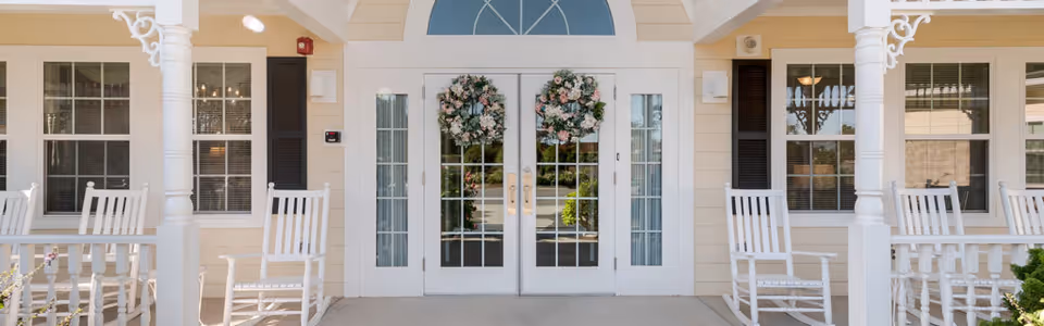 Front entrance of a senior living facility with double glass doors decorated with floral wreaths, flanked by windows and white rocking chairs on a covered porch.