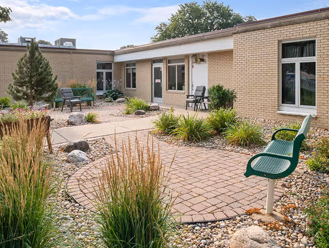 Outdoor courtyard with a brick-paved seating area, green benches and chairs, and landscaped plants in front of a single-story brick building.
