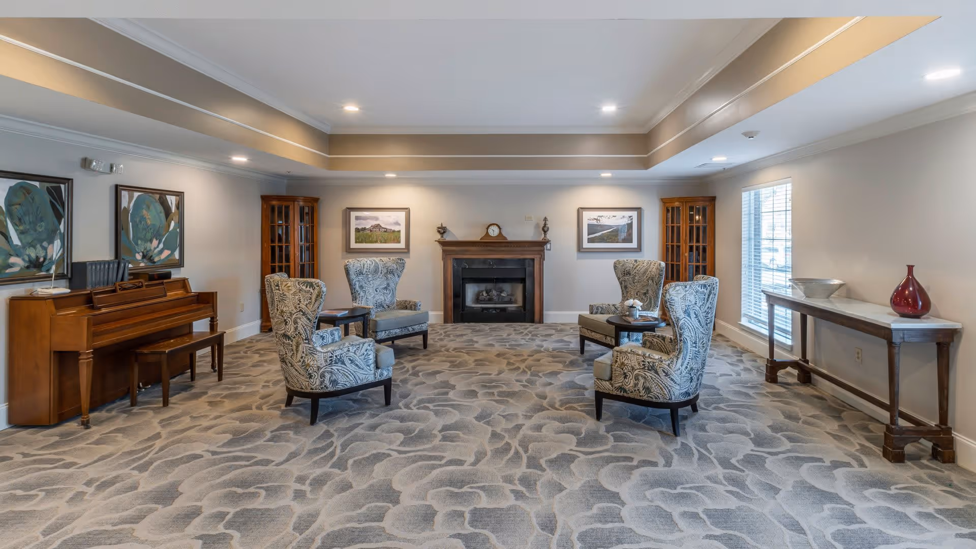 A spacious living room with patterned carpet, four upholstered armchairs arranged around two small tables, a wooden piano with a bench, two glass-fronted wooden cabinets, a fireplace with a clock and decorative items on the mantel, framed artwork on the walls, and a long console table with decorative bowls and a vase near a window with blinds.