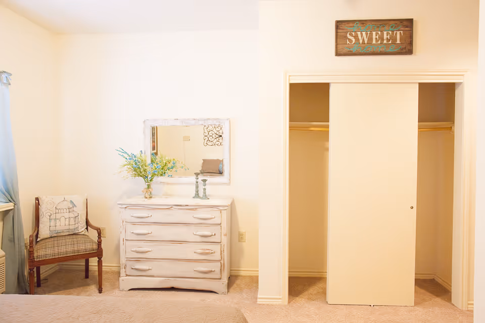 A cozy bedroom corner with a wooden chair featuring a decorative pillow, a white distressed dresser with a vase of flowers and two candle holders on top, a wall mirror above the dresser, and a closet with sliding doors. Above the closet is a wooden sign that reads 'home SWEET home'.