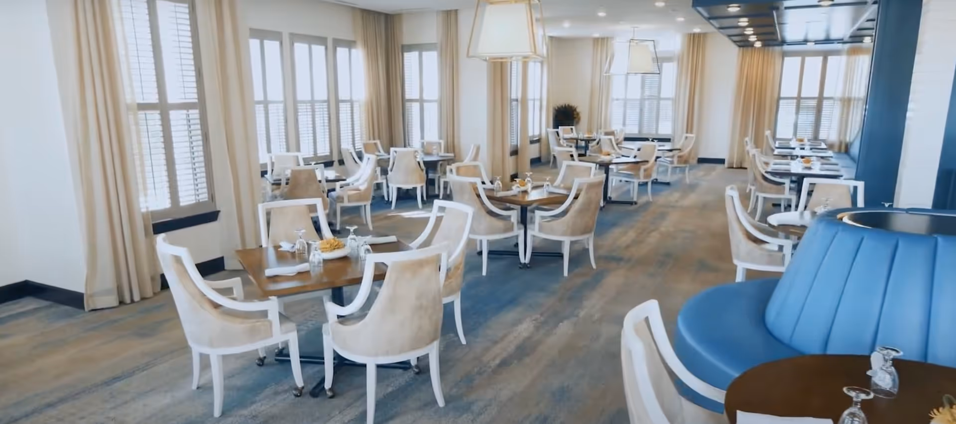 A bright and spacious dining room with multiple wooden tables and beige upholstered chairs arranged neatly. Large windows with white shutters and beige curtains allow natural light to fill the room. Modern pendant lights hang from the ceiling, and a blue cushioned circular seating area is visible in the foreground.