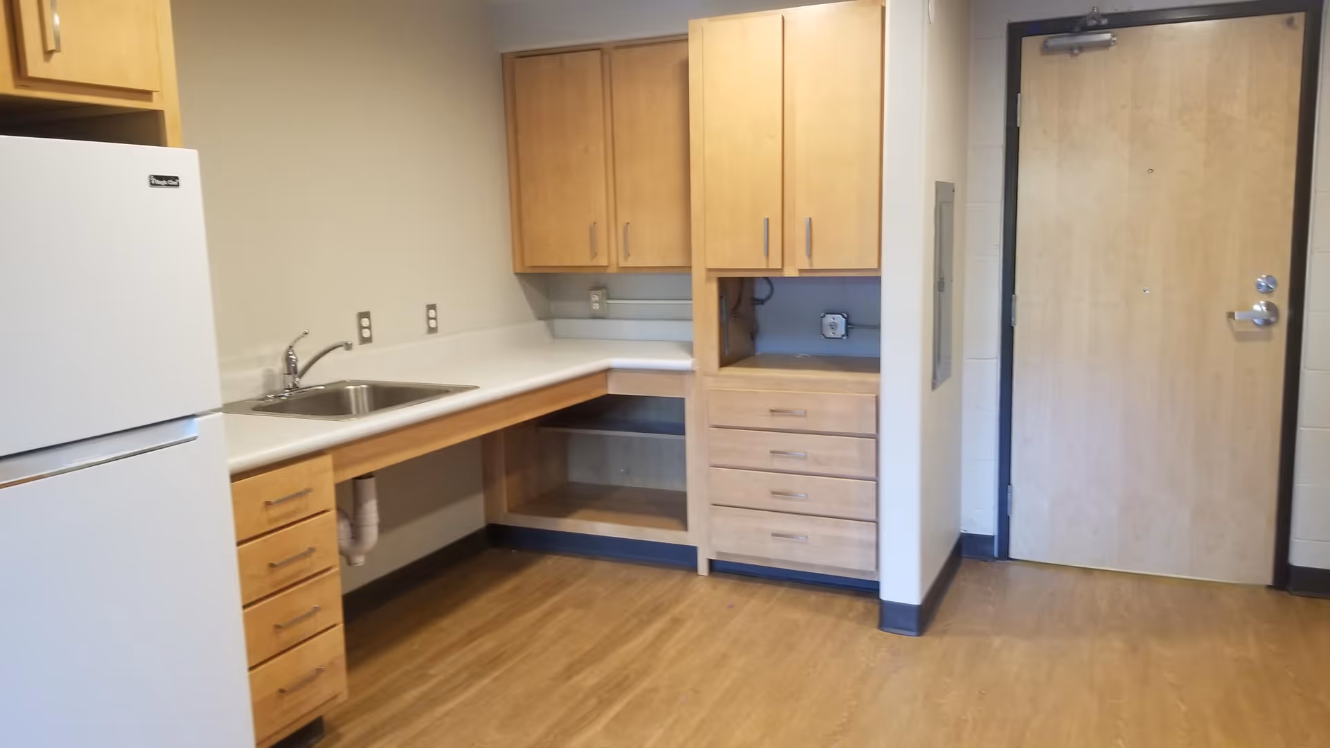 Kitchenette area with a refrigerator, sink, light wood cabinets and drawers, and a wooden door.