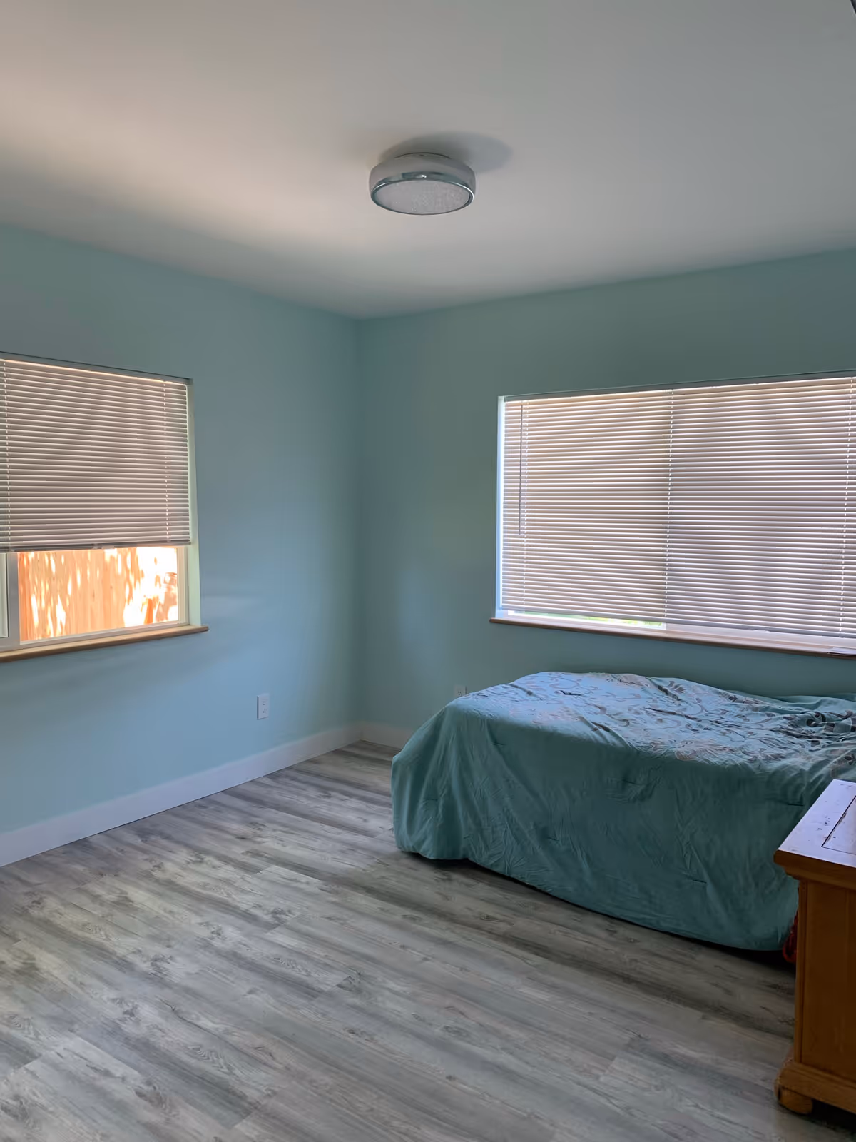 A simple bedroom with light blue walls and two windows with closed blinds. There is a single bed with a blue-green bedspread positioned against one wall and a wooden nightstand next to it. The floor is covered with light-colored wood laminate, and a ceiling light fixture is visible.