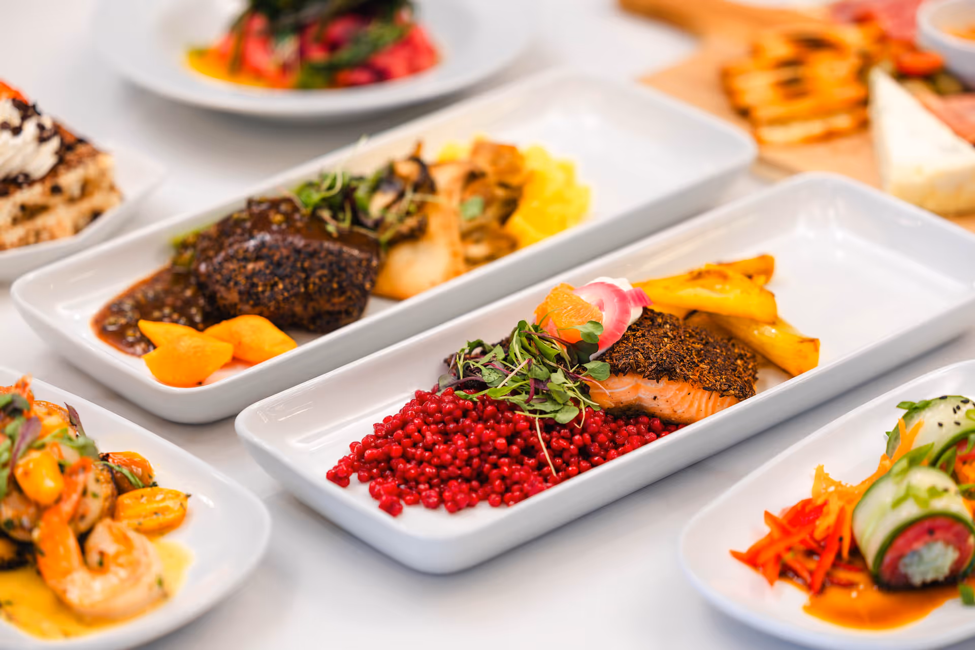 Close-up view of several plated gourmet dishes including a herb-crusted salmon with red berries and roasted vegetables, shrimp with yellow cherry tomatoes, and a sushi roll with cucumber and carrot garnish, all served on white rectangular plates.