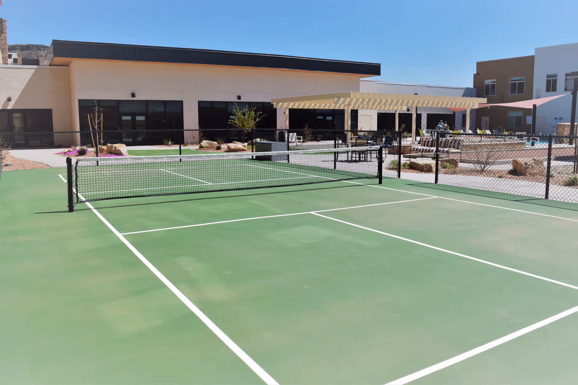 Outdoor tennis or pickleball court with a net in the foreground, surrounded by a black chain-link fence. In the background, there is a modern building with large windows, a shaded seating area with tables and chairs, and some landscaping with rocks and plants under a clear blue sky.