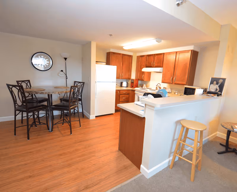 Interior view of a senior living facility kitchen and dining area. The kitchen features wooden cabinets, a white refrigerator, stove, microwave, and a counter with a wooden stool. The dining area has a round table with four chairs and a wall clock above it. The floor is a combination of wood and carpet.
