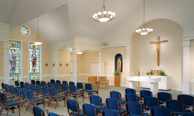 Interior chapel with rows of blue chairs facing an altar, crucifix, stained glass windows and religious statues.