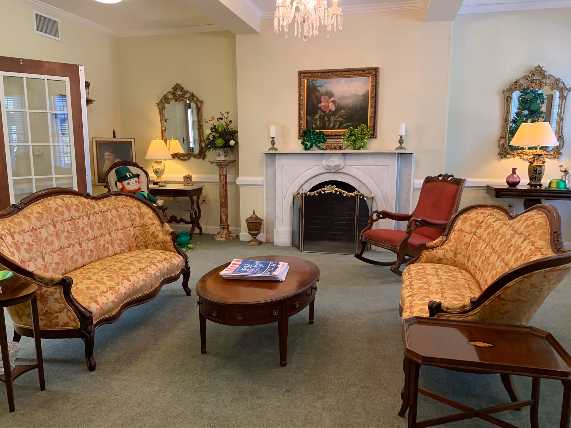 A cozy living room with two vintage floral-patterned sofas facing each other, a wooden coffee table with magazines on top, a red upholstered rocking chair, and a white marble fireplace decorated with green shamrocks and candles. The room also features ornate mirrors, a chandelier, a side table with a lamp, and a leprechaun doll sitting on one of the sofas.