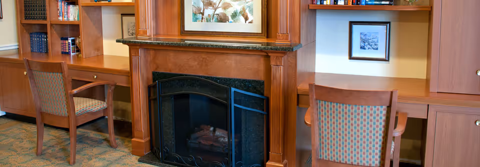 A cozy interior room featuring a wooden fireplace with a black metal screen, flanked by built-in wooden desks and shelves. Two wooden chairs with patterned upholstery are placed at the desks. Books and framed artwork decorate the shelves and walls.