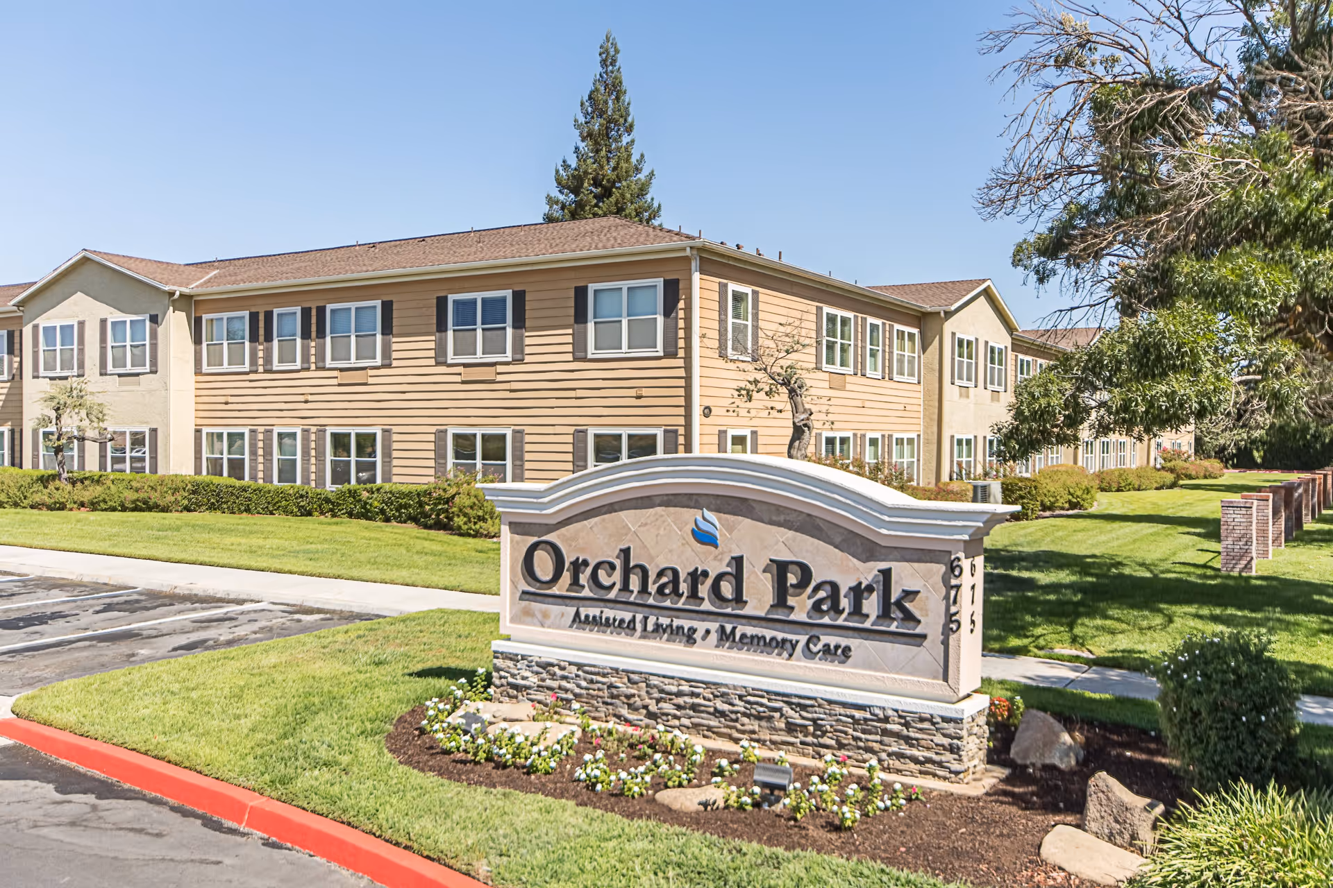 Exterior view of a senior living facility named Orchard Park with a large sign in front that reads 'Orchard Park Assisted Living / Memory Care'. The building is two stories with beige and light brown siding, multiple windows, and surrounded by well-maintained green lawns and trees under a clear blue sky.