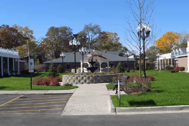 Outdoor view of a senior living facility courtyard with a paved walkway leading to a central stone fountain surrounded by green grass, bushes, and lamp posts. Buildings with white railings and columns flank the courtyard on both sides under a clear blue sky.