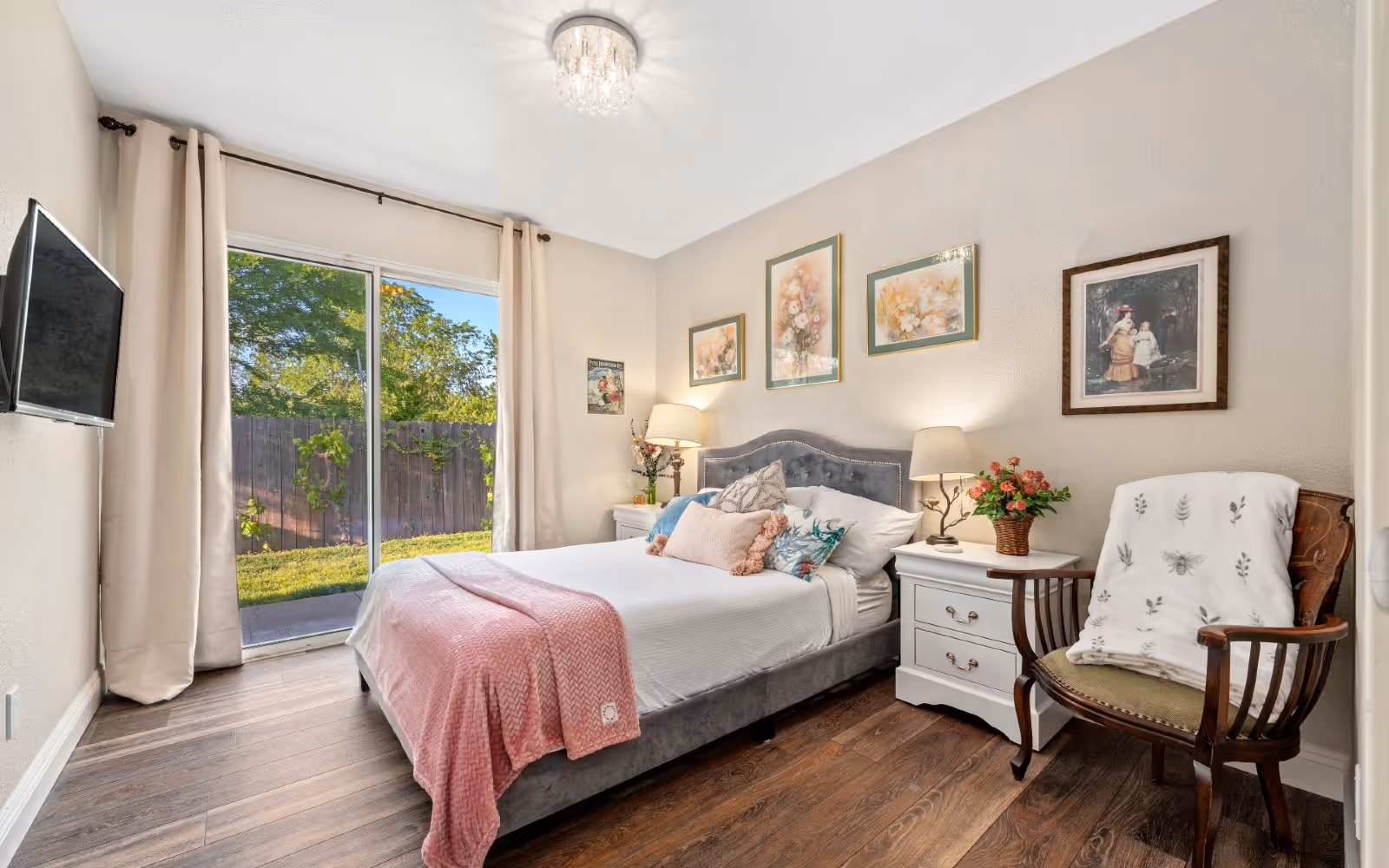 Bright furnished bedroom with a bed, nightstands, framed artwork, an armchair, and sliding glass doors overlooking a yard.
