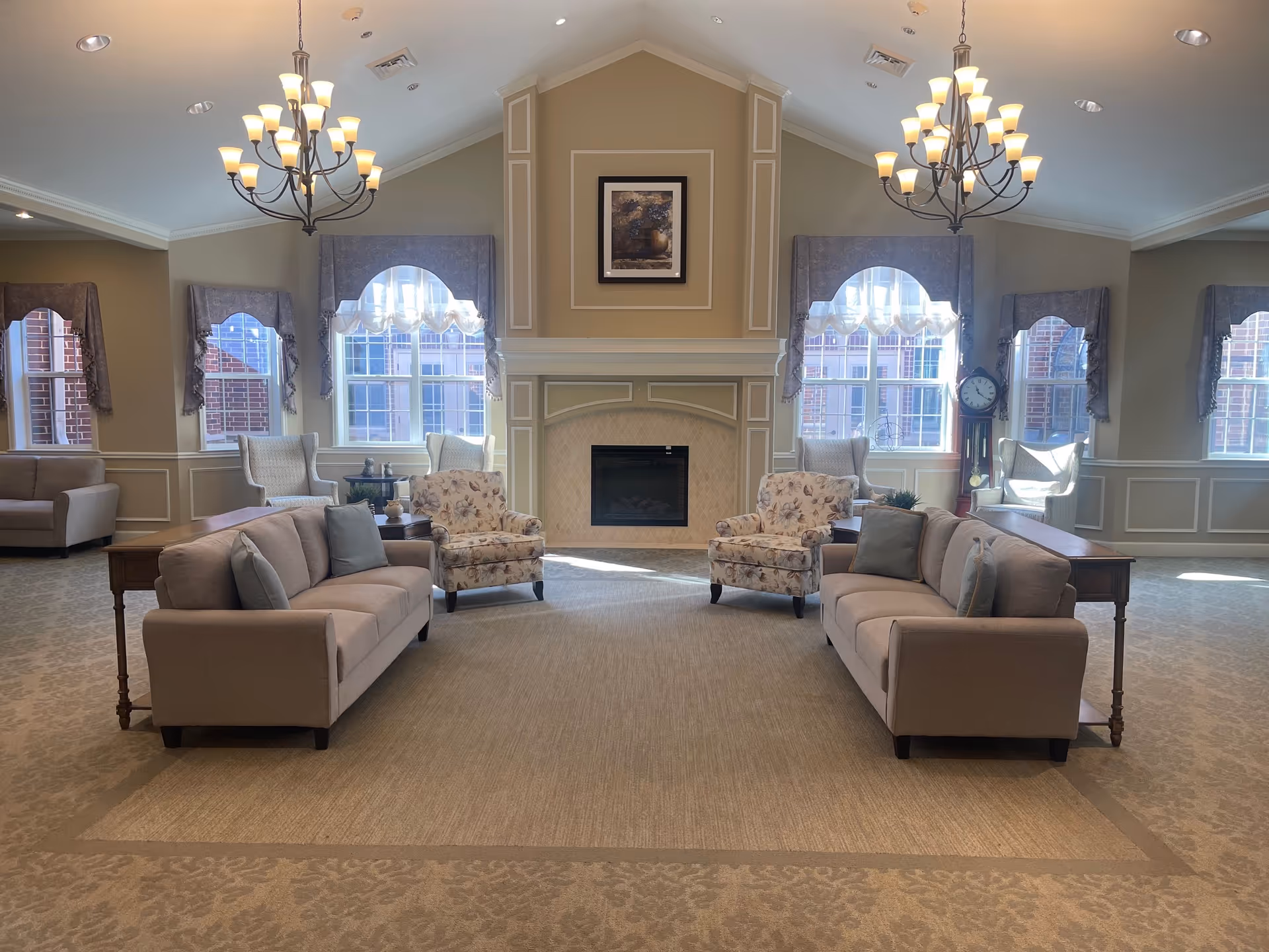 A spacious and well-lit living room area with two beige sofas facing each other, two floral armchairs, and two white wingback chairs near large windows with decorative curtains. The room features a central fireplace with a framed picture above it, two large chandeliers hanging from a vaulted ceiling, and a grandfather clock near the windows.