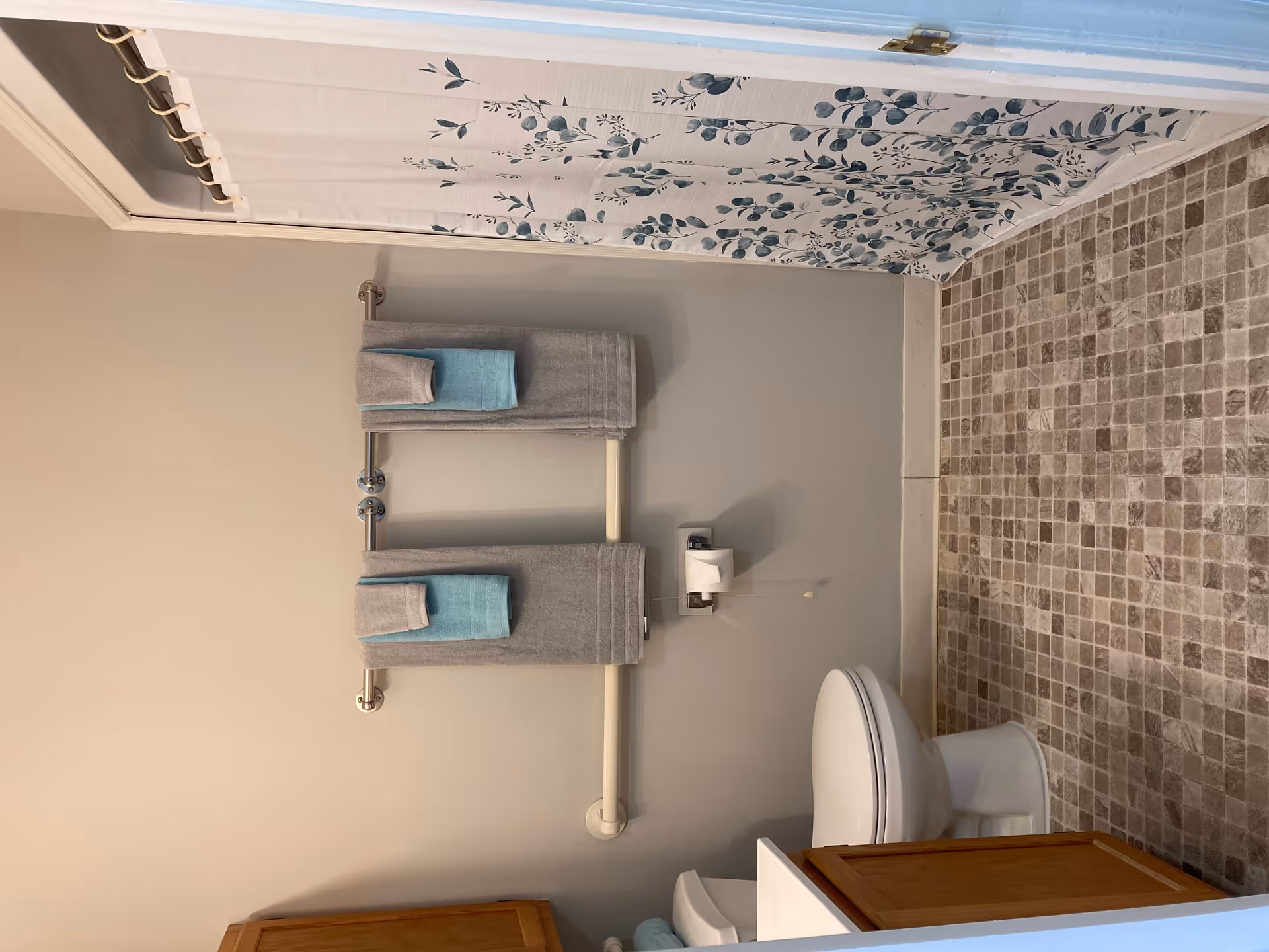 A bathroom with a tiled floor, a toilet, a wooden cabinet, and a shower with a floral-patterned curtain. Two towel racks hold gray and blue towels on the wall above a grab bar.