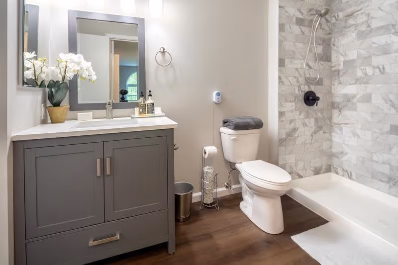 A modern bathroom featuring a gray vanity with a white countertop, a large mirror above it, and a potted white orchid on the counter. Next to the vanity is a white toilet with a folded gray towel on top, a toilet paper holder with extra rolls, and a small trash can. The shower area has a tiled wall with a mounted showerhead and a white shower base. The floor is wood-style, and a white bath mat is placed in front of the shower.