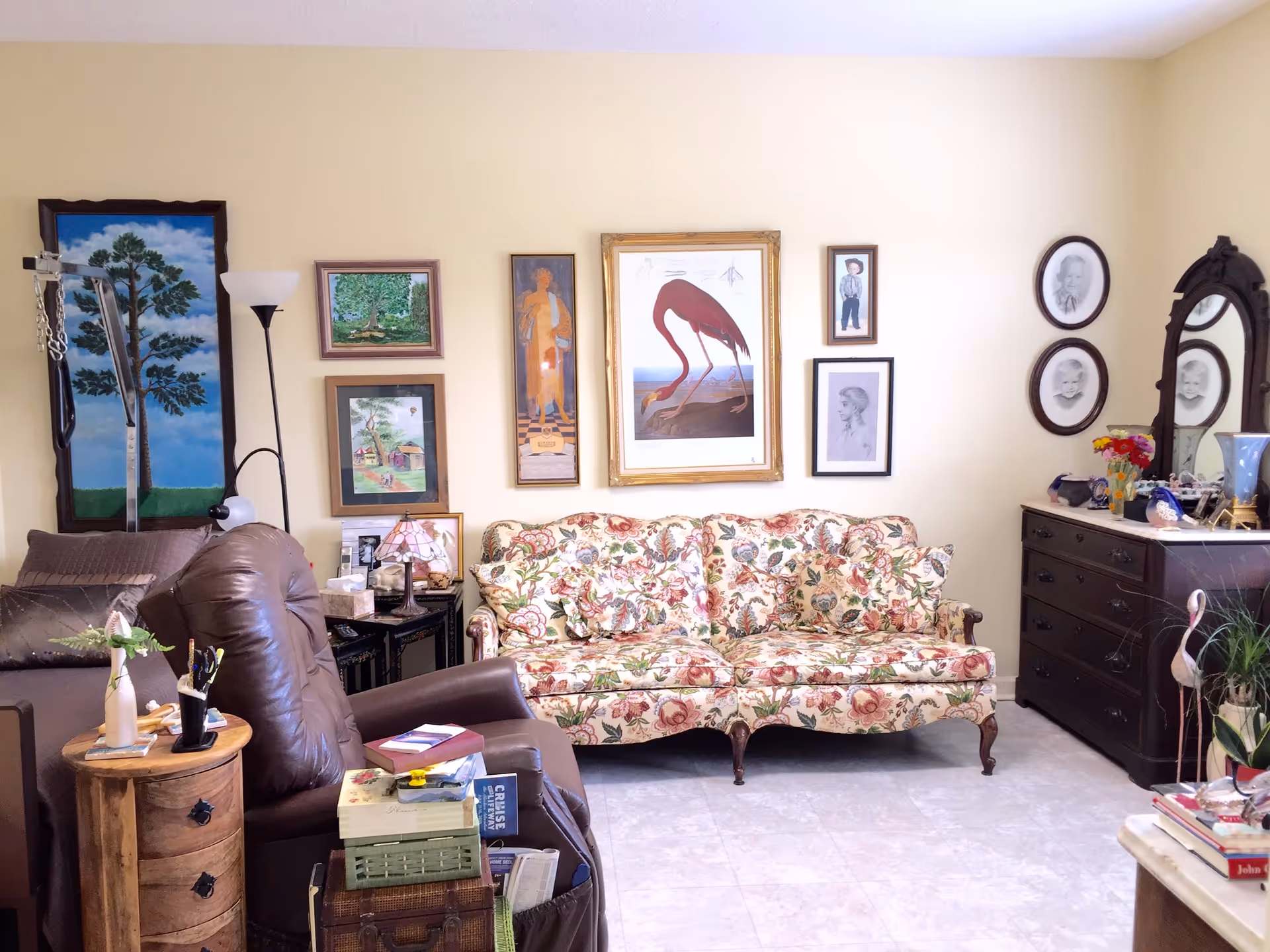 A cozy living room with a floral patterned sofa, a brown leather recliner chair, and a wooden side table with books and decorative items. The walls are adorned with various framed paintings and portraits. There is a tall floor lamp next to the recliner and a dark wooden dresser with a mirror and more decorative items on the right side.