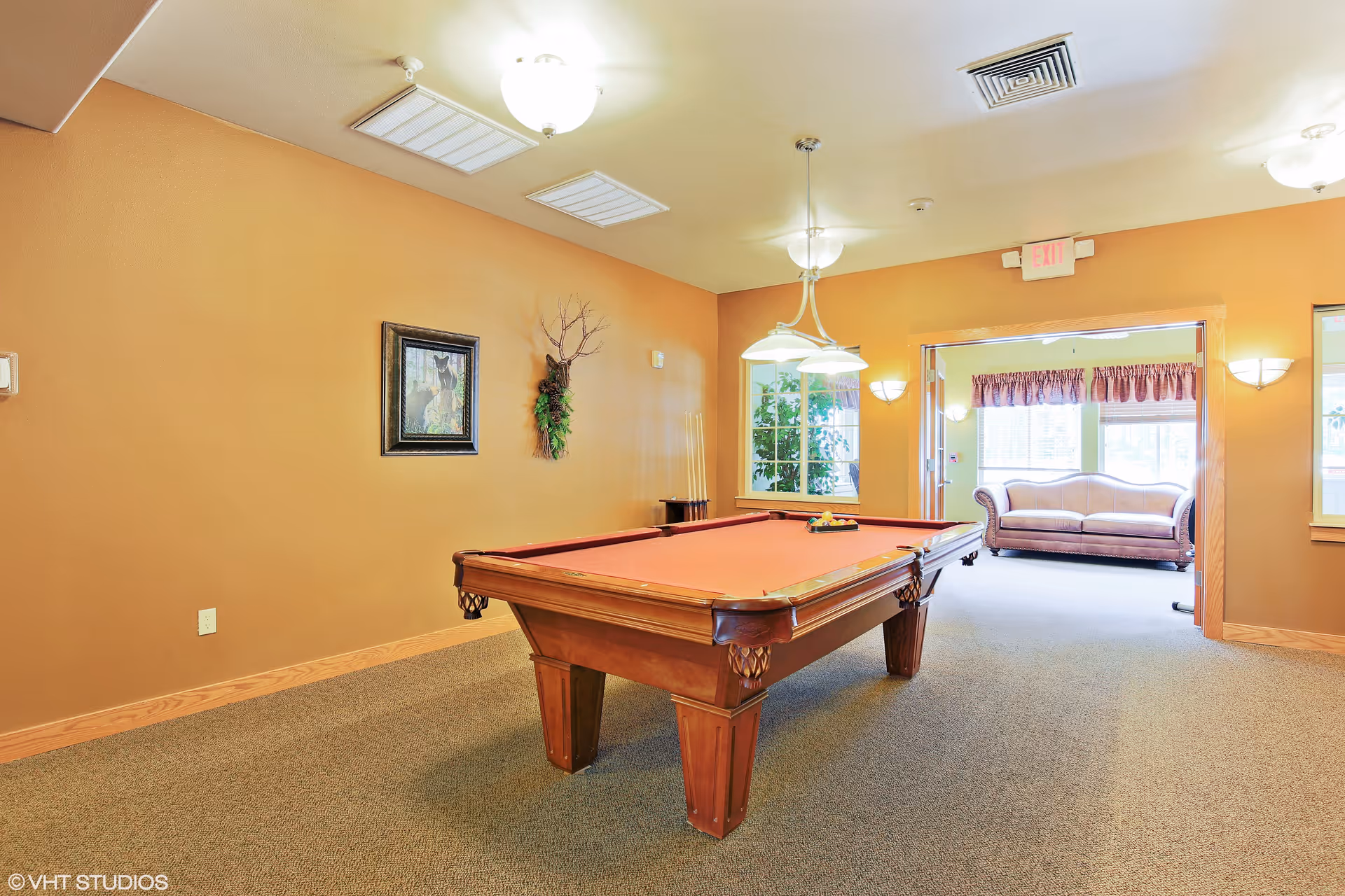 A well-lit recreational room with a pool table in the center. The walls are painted a warm beige color and decorated with a framed picture and a wall-mounted plant. There is a window with a view of greenery and a doorway leading to a bright sitting area with a sofa and windows with valances.