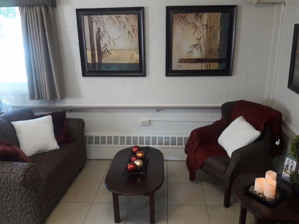 A cozy living room area with a patterned sofa and an armchair, both with white pillows. The armchair has a red throw blanket draped over it. A small wooden coffee table with decorative red and gold balls is placed between the seating. Two framed paintings with abstract bamboo designs hang on the white brick wall behind the furniture. A side table with three lit candles and a small plant is visible on the right side.