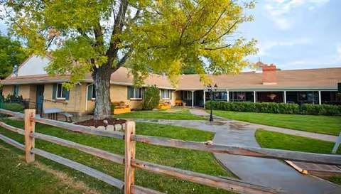 Single-story senior living facility exterior with a wooden fence, tree, and green lawn.