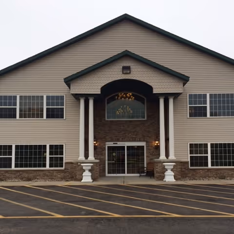 Front exterior view of a two-story building with beige siding and stone accents. The entrance features a covered porch supported by four white columns and a glass door. Multiple large windows are visible on both floors. The parking area in front has yellow-striped lines.