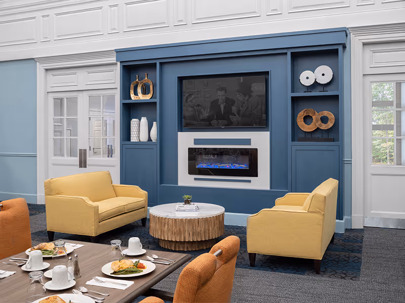 A cozy living room area in a senior living facility with two yellow armchairs facing a blue built-in wall unit that houses a flat-screen TV and an electric fireplace. The wall unit also features decorative shelves with vases and art pieces. In the foreground, there is a dining table set with plates of food, cups, and utensils, surrounded by orange upholstered chairs. The room has light blue walls with white trim and large windows letting in natural light.